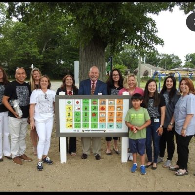 Smiling teachers, therapists, administrators and students standing around a newly installed playground communication board