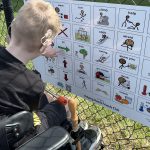 young man with cochlear implant pointing to a message on a playground communication board.